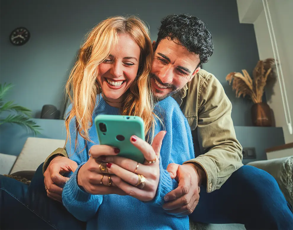 Pareja en el sofá sonriendo al smartphone, en un acogedor ambiente hogareño.