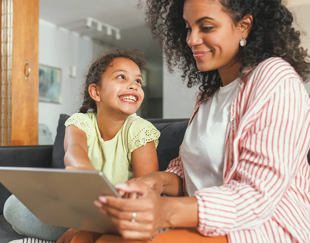 Mother and daughter sitting on a couch using a tablet together in a cozy living room, highlighting family-focused cybersecurity awareness.