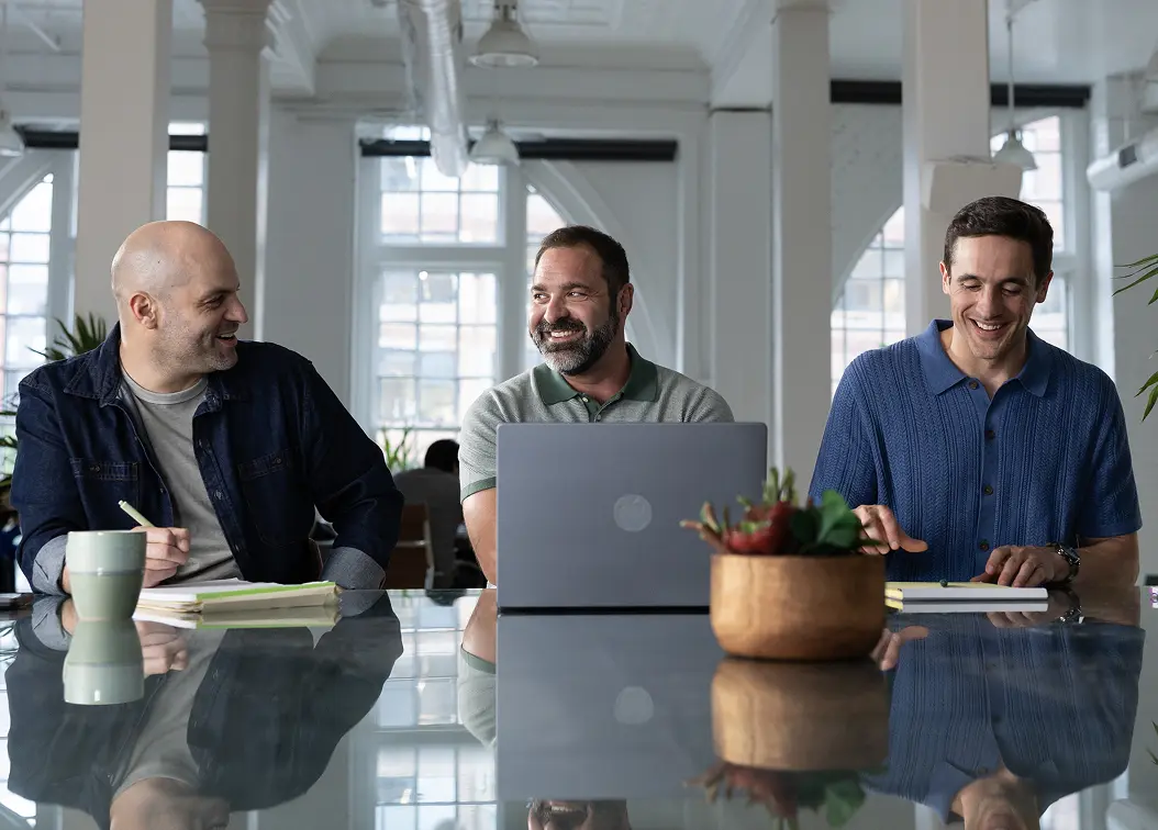 Three male business partners sitting around a table during a meeting, smiling and engaged in conversation.