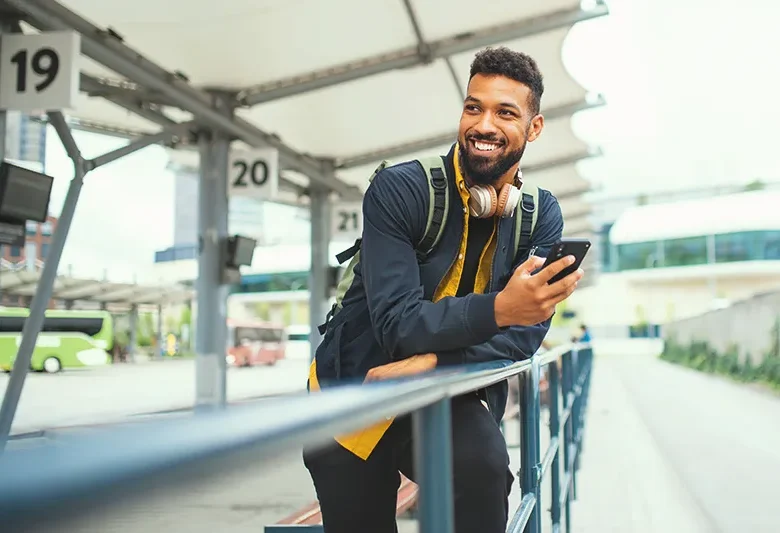 Traveler with backpack checking phone at a bus terminal with numbered platforms and buses in the background