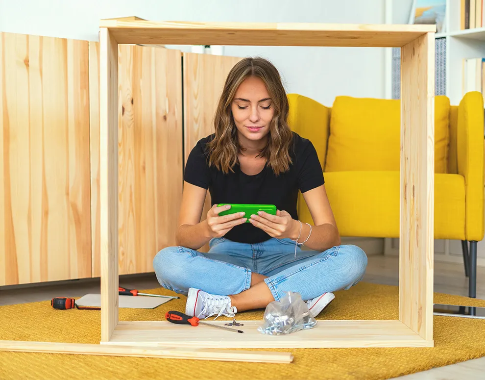 Person sitting on the floor assembling furniture while using a smartphone, representing eBranch support and digital self service tools