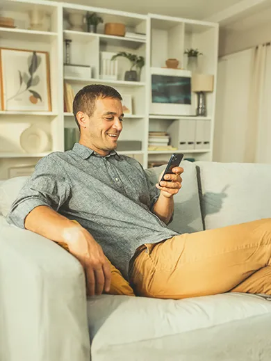 Man sitting on a couch at home, smiling as he looks at his phone.